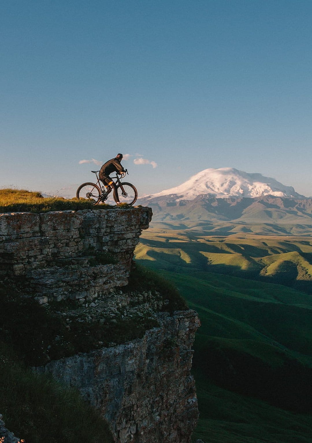 Mountain biker standing on a rocky precipice overlooking a green valley and a massive snow-capped peak under blue sky