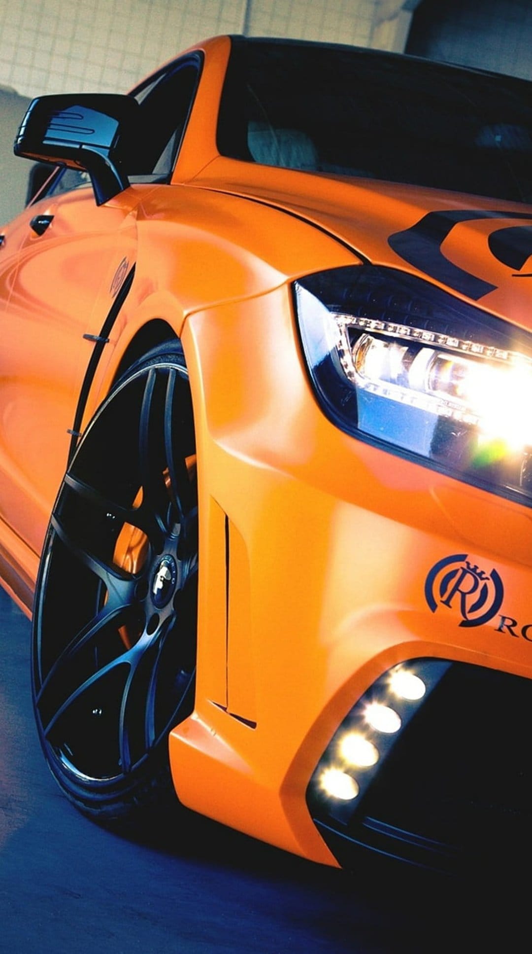 Orange sports car showcases custom black rims, red brake calipers, and glowing LED headlights in a close-up.