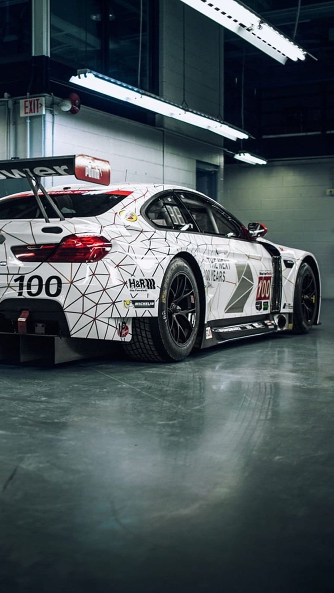 BMW M4 DTM race car with geometric patterns and a large rear wing parked in a brightly lit indoor garage area.