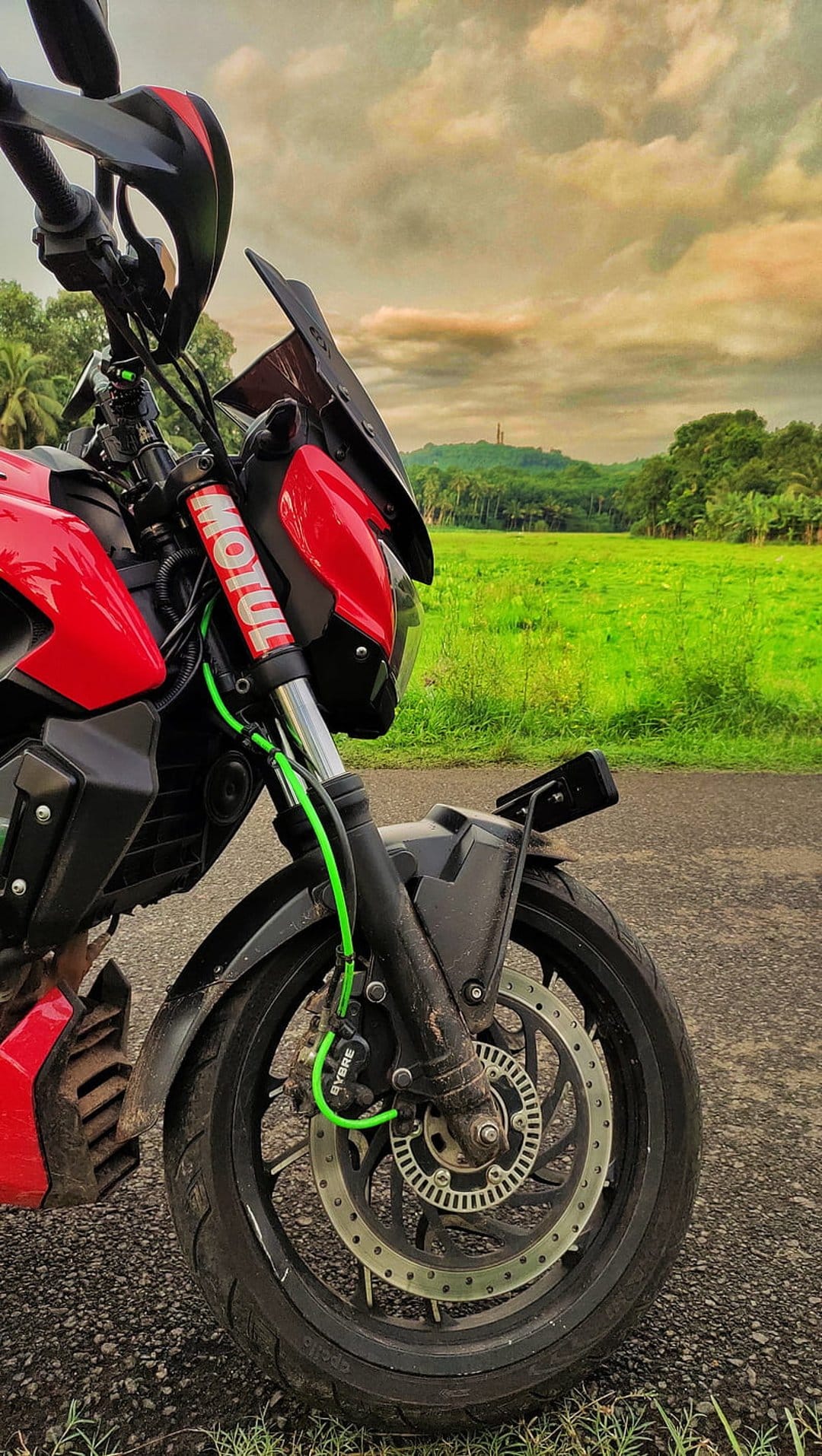 Red motorcycle front wheel and fairing on a gravel road with green hills under a cloudy sky in the background.