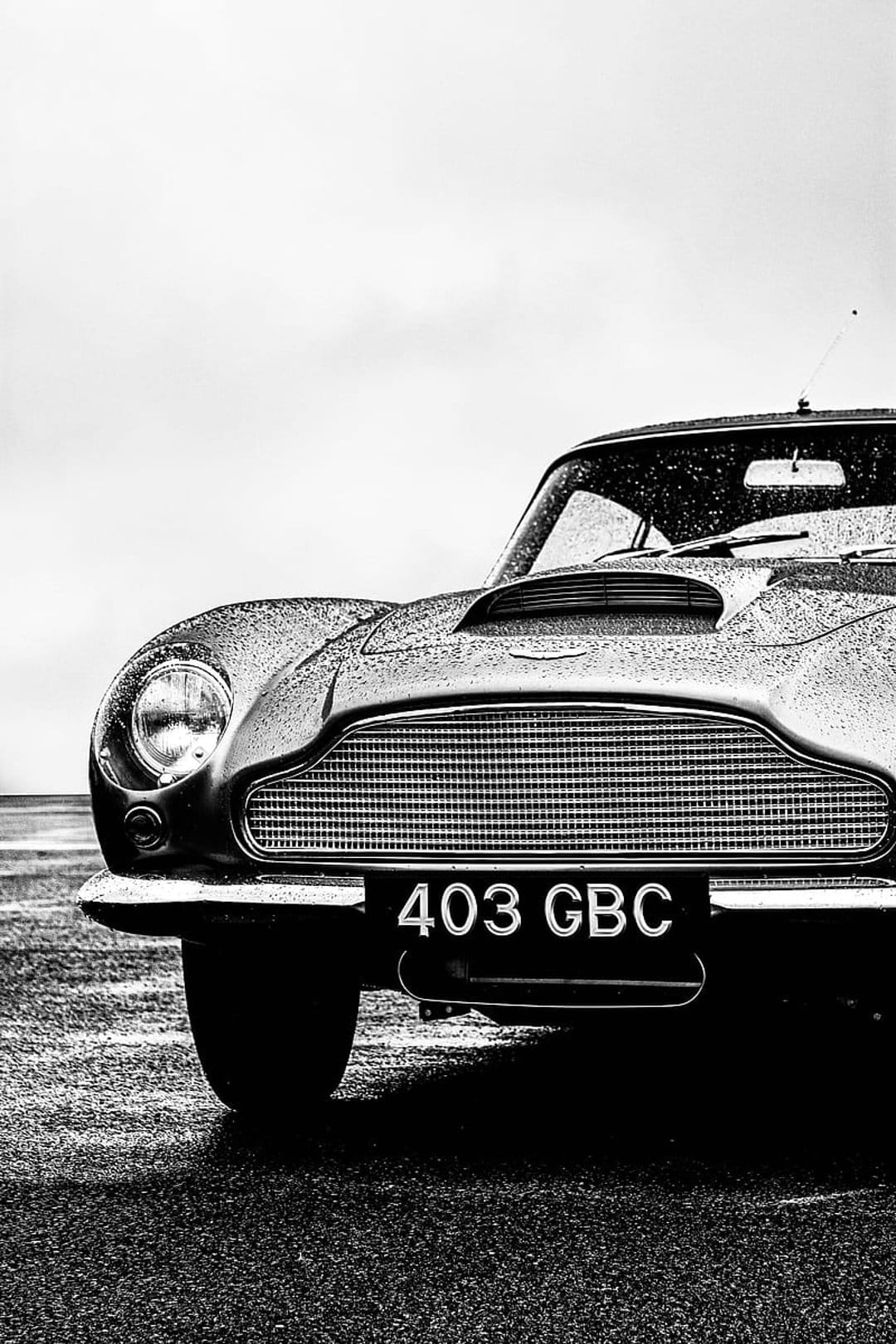Aston Martin DB5 front grille and headlights covered in raindrops during a moody black and white rainstorm.