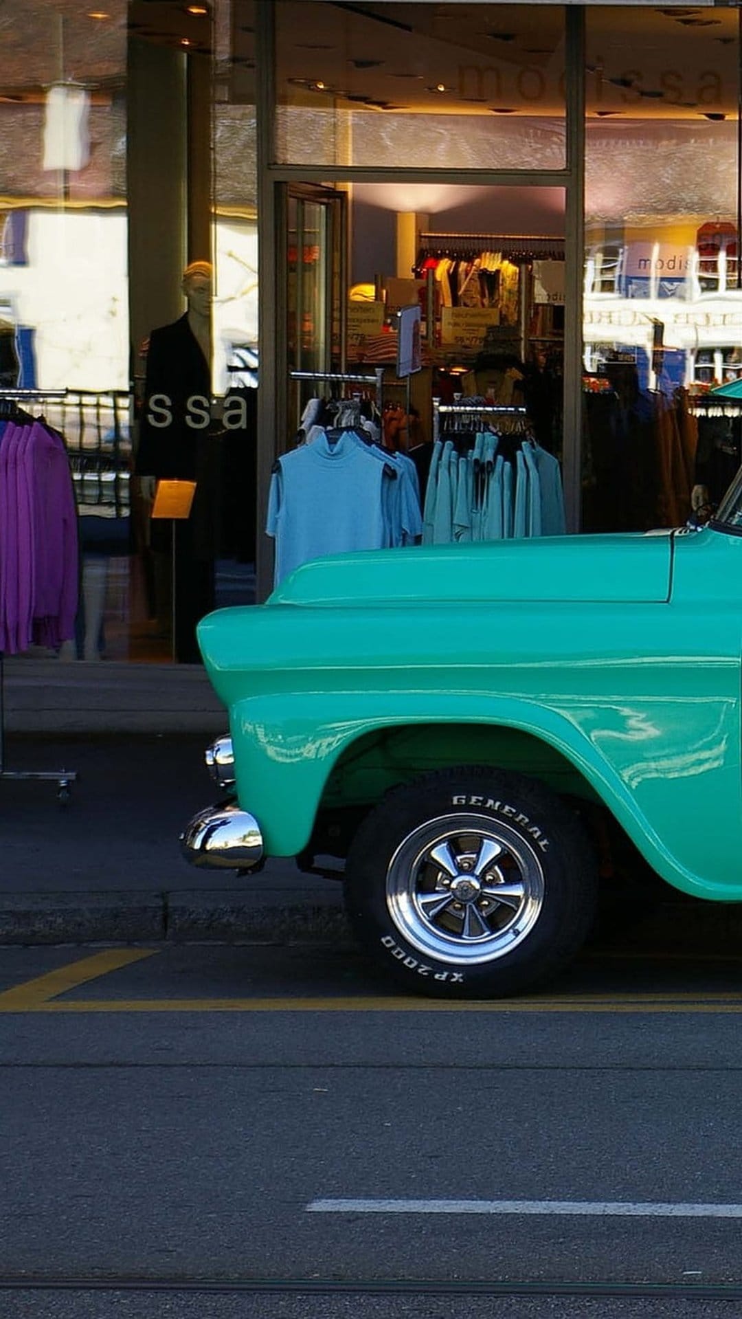 Teal vintage truck with chrome accents parked in front of a boutique window featuring clothing and mannequins.