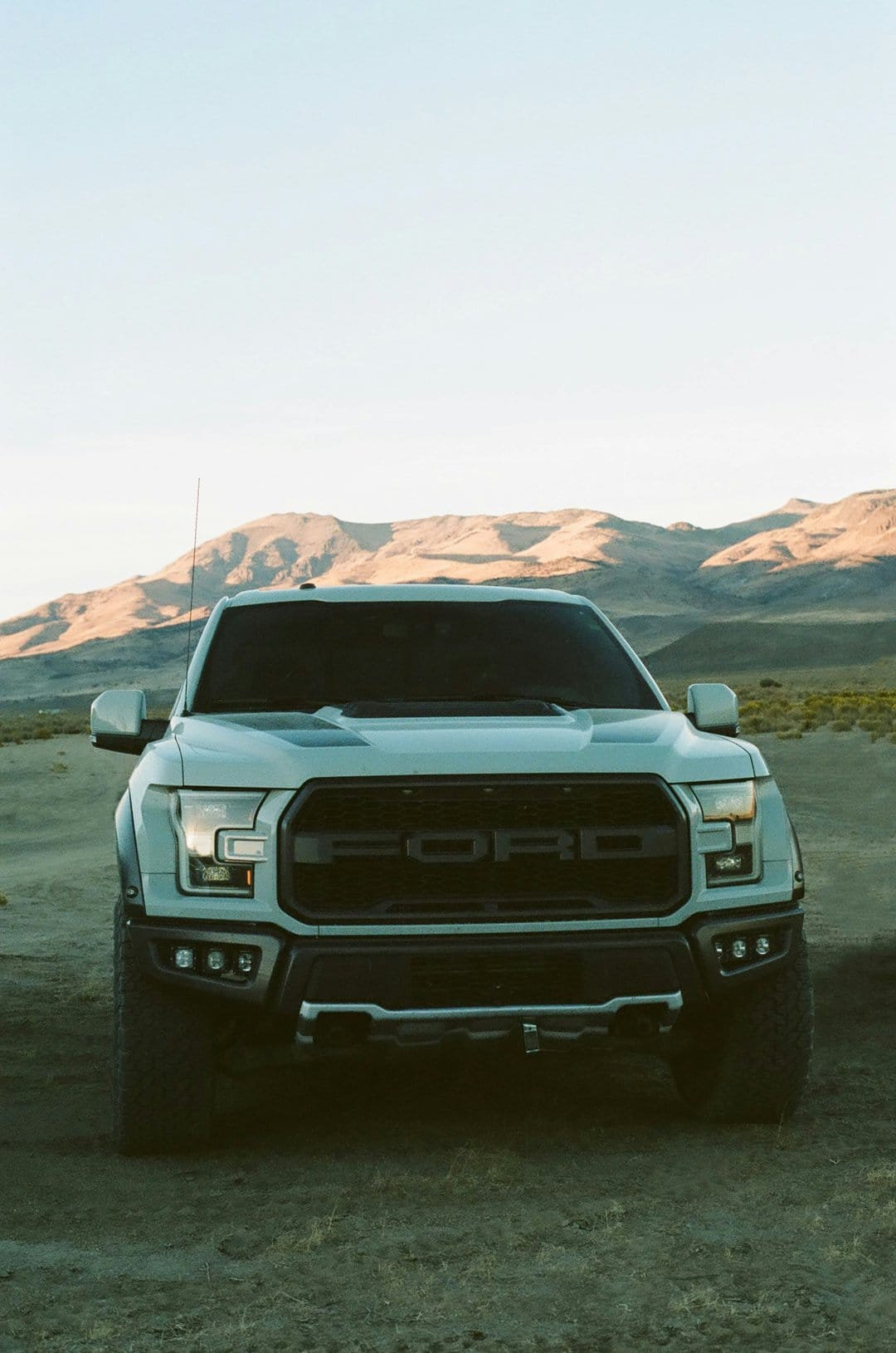White Ford Raptor truck with a black grille and LED lights parked on a desert road during golden hour.
