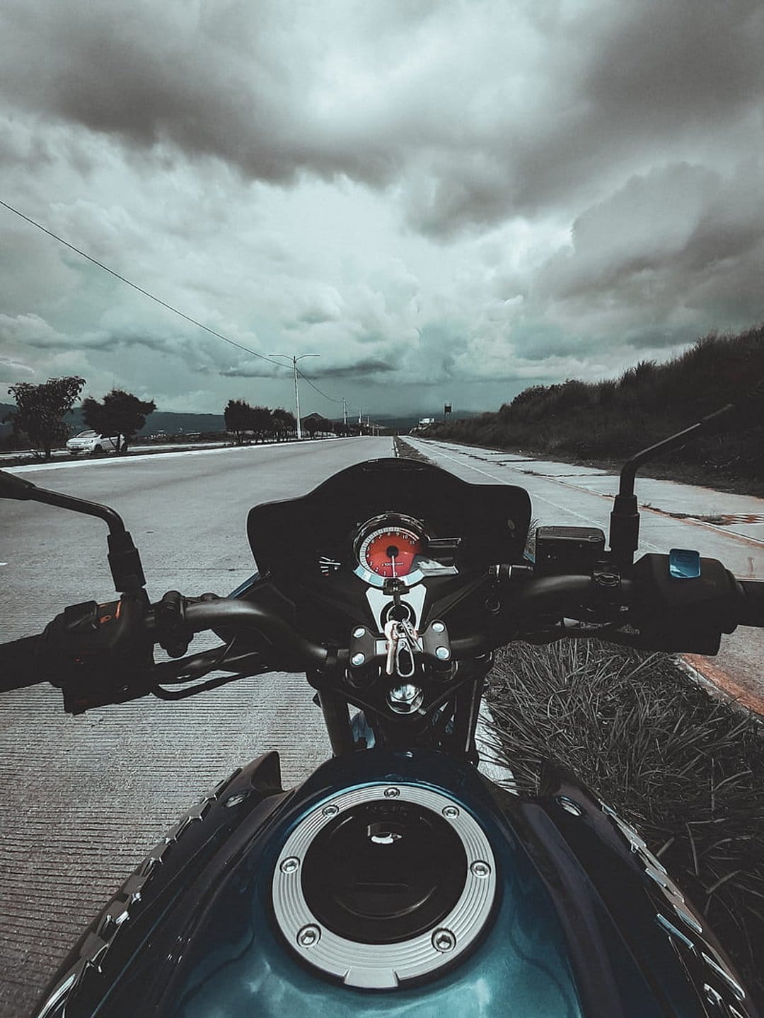 Motorcycle handlebars and a red-lit speedometer face a dark, stormy sky on an empty rural road during a journey.