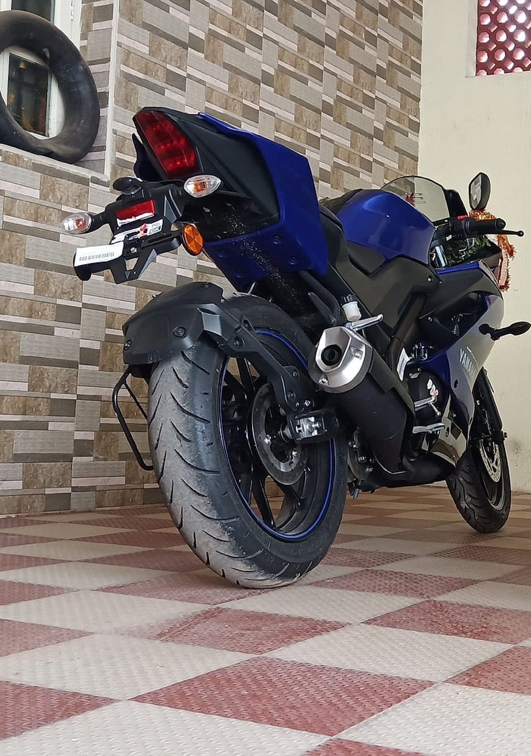 Blue Yamaha R15 motorcycle parked on a checkered floor with its rear tire and exhaust visible against a brick wall.