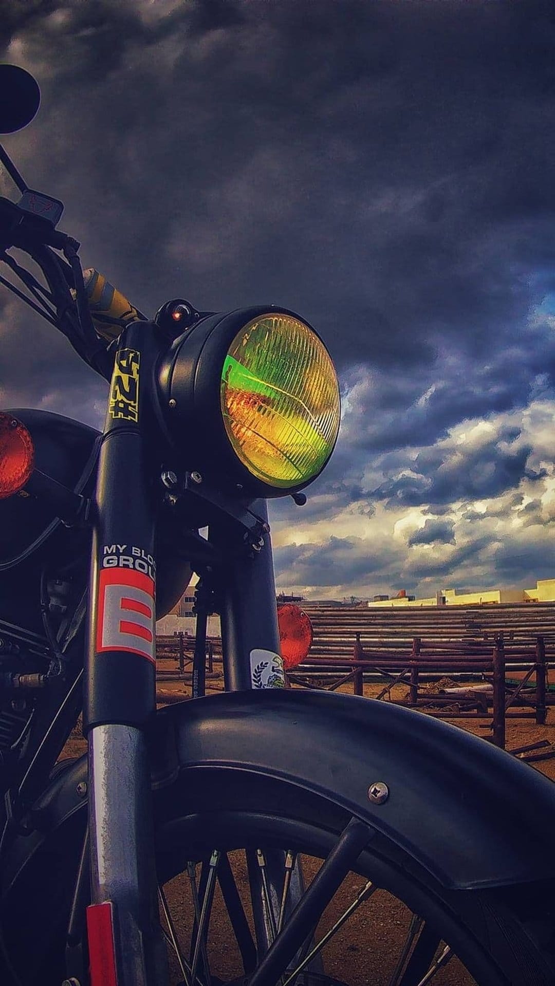 Vintage motorcycle headlight with yellow lens reflecting dark clouds, chrome forks, and black metal frame.
