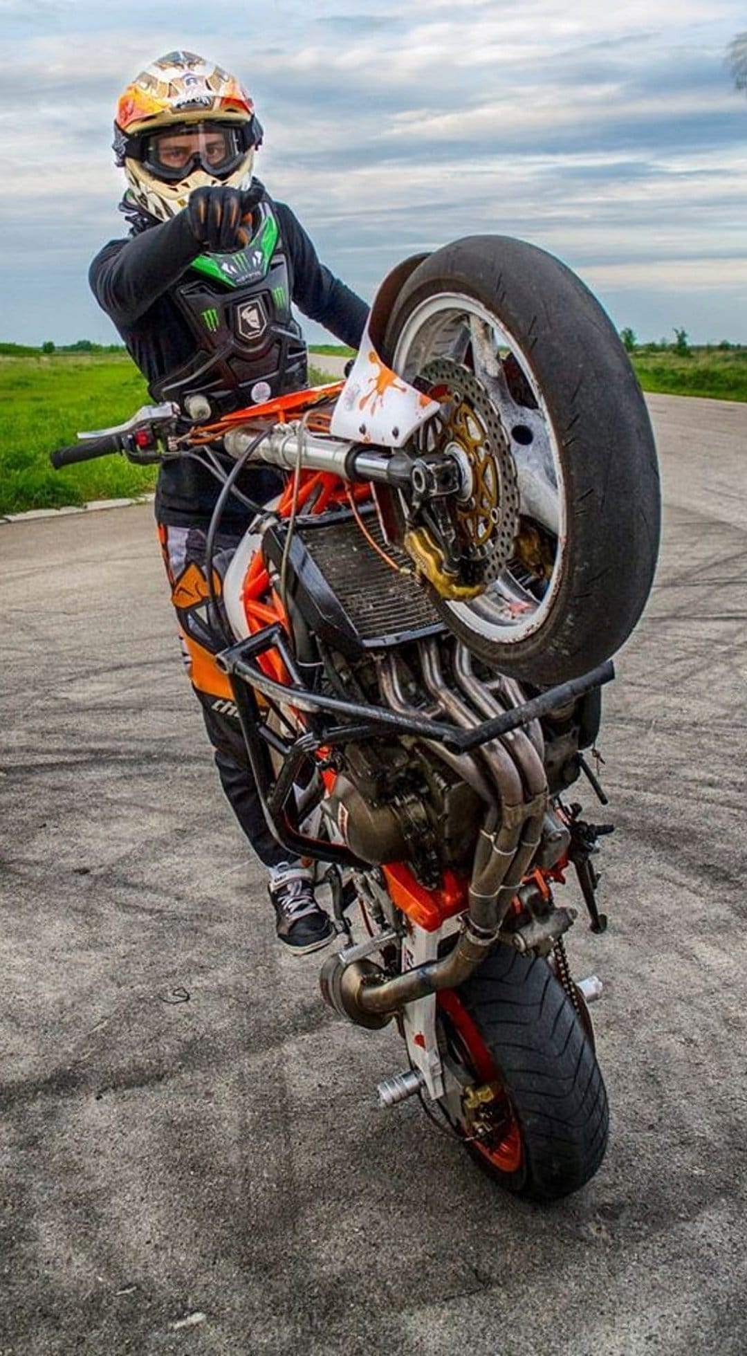 Motorcyclist in full armor performs a vertical wheelie showing an exposed engine against a cloudy gray sky.