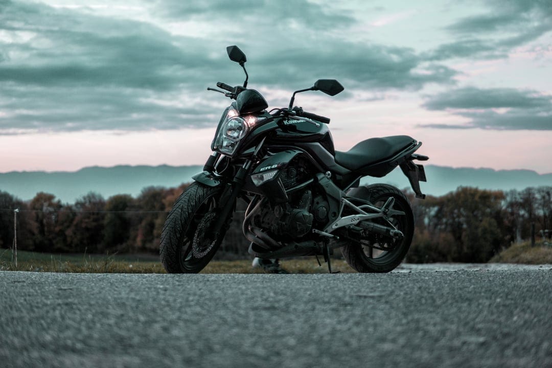 Matte black motorcycle parked on asphalt under a dramatic orange sunset sky with silhouettes of distant hills.
