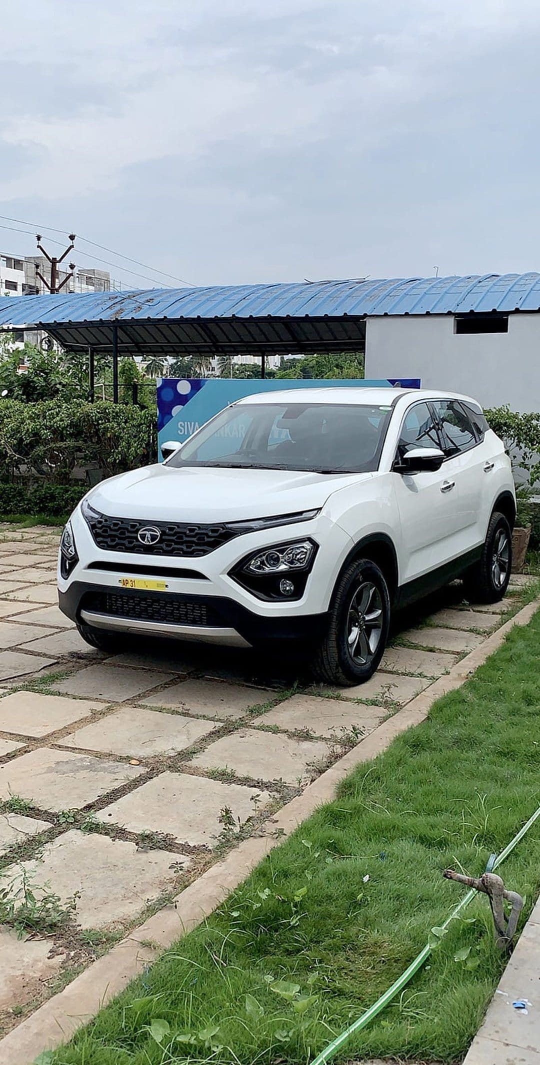 White Tata Harrier SUV parked on a square concrete driveway with green grass borders under a cloudy sky.