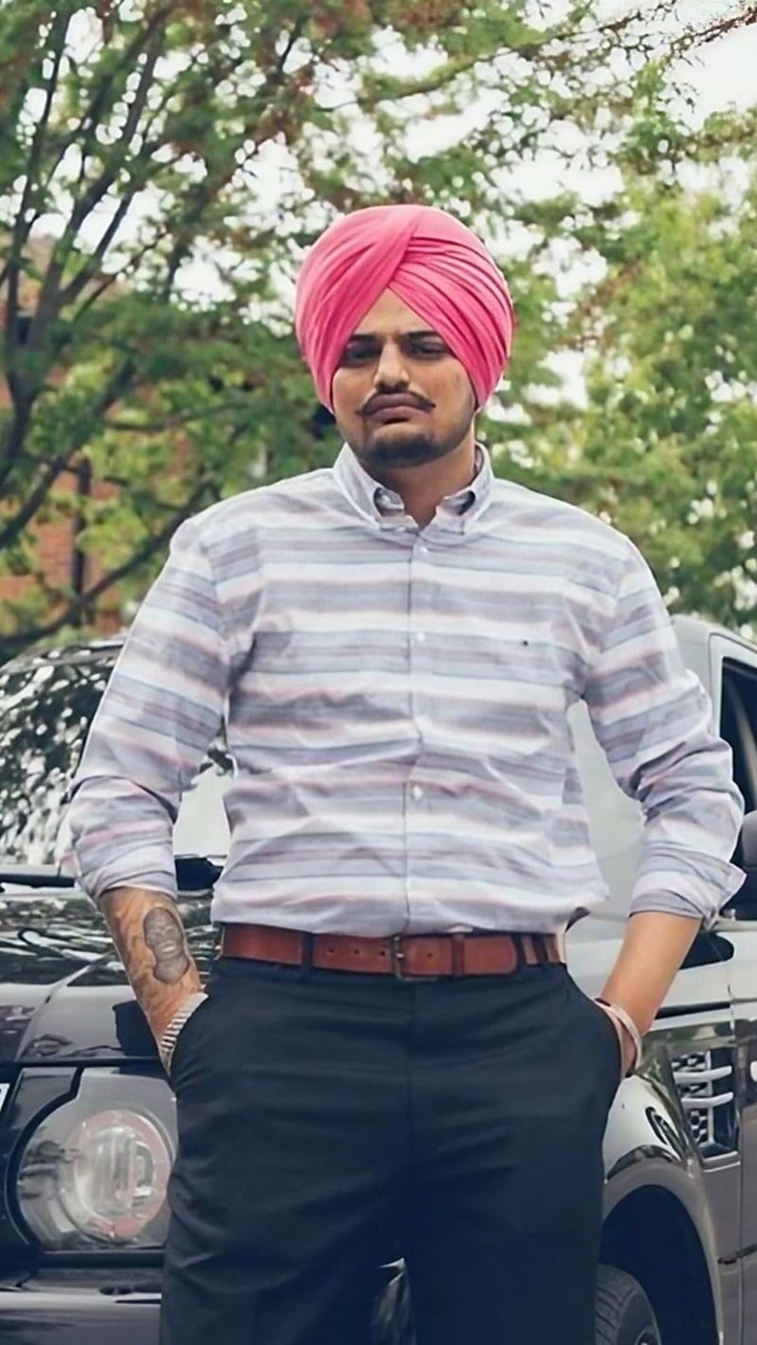 Sidhu Moose Wala in a pink turban and striped shirt leans against a dark SUV with trees in the background.