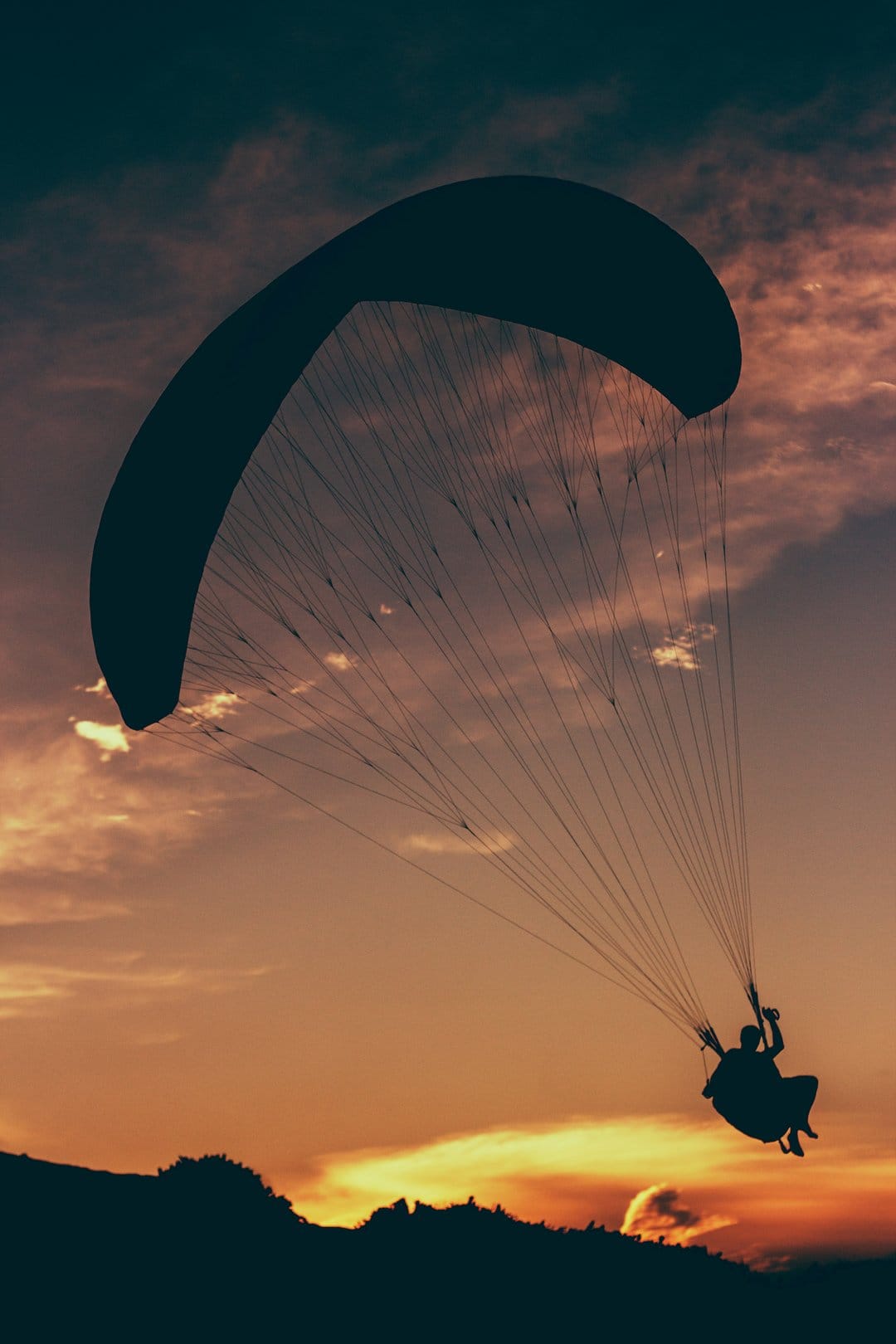Paraglider pilot silhouetted against orange and pink sunset clouds above dark rolling hills in a vertical view.