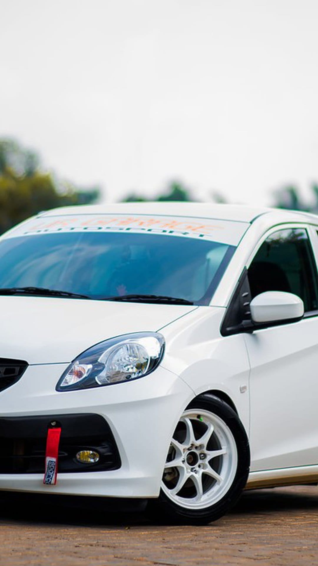 White Honda Brio hatchback with an orange sun visor, red tow hook, and multi-spoke wheels in an outdoor setting.