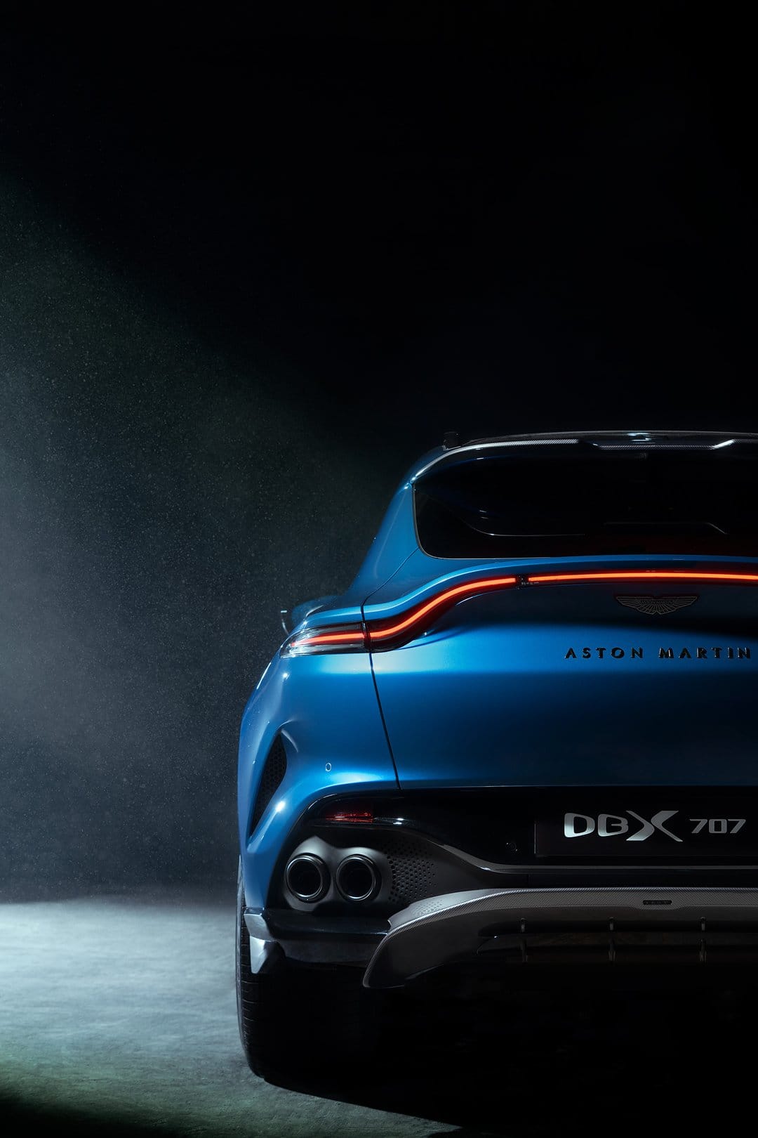 Rear view of a blue Aston Martin DBX 707 with a glowing red taillight bar in a dark, atmospheric setting.