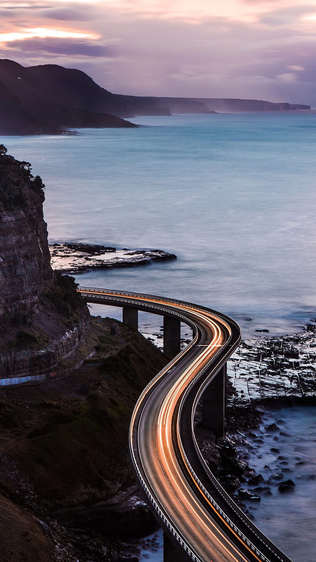 Winding coastal bridge with glowing light trails hugging rocky cliffs above a calm sea during late twilight hours.