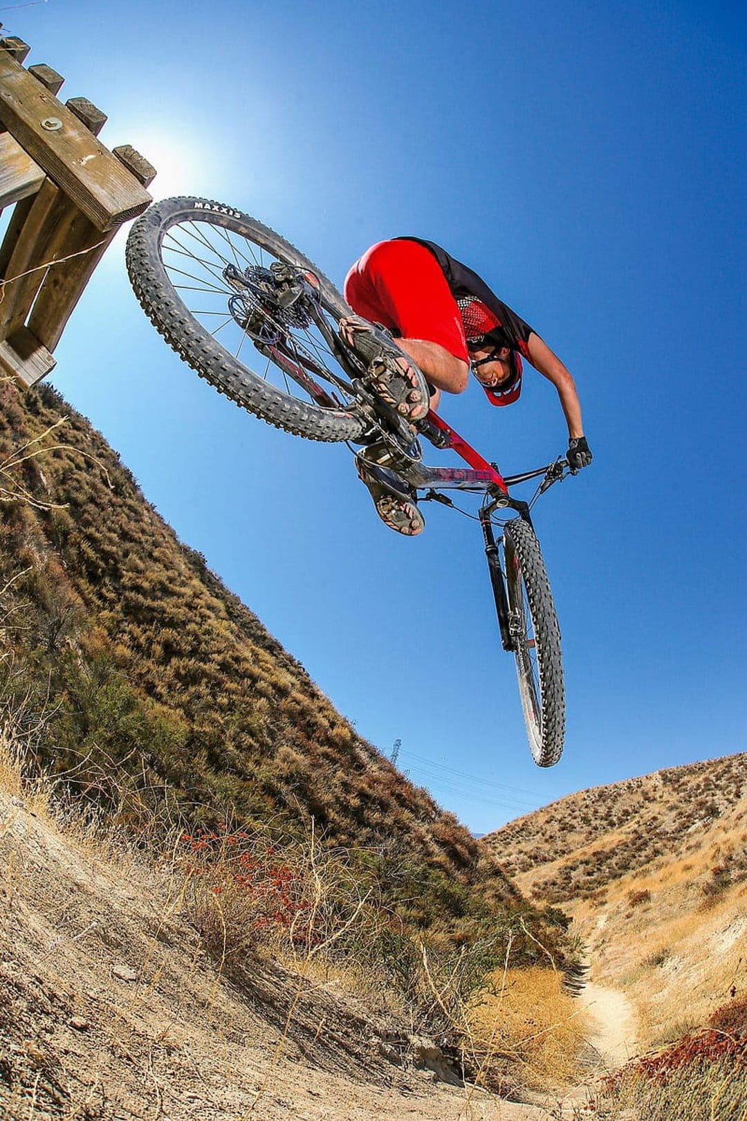 Mountain biker in red shorts jumps a bicycle off a wooden ramp into a clear blue sky over hilly terrain.