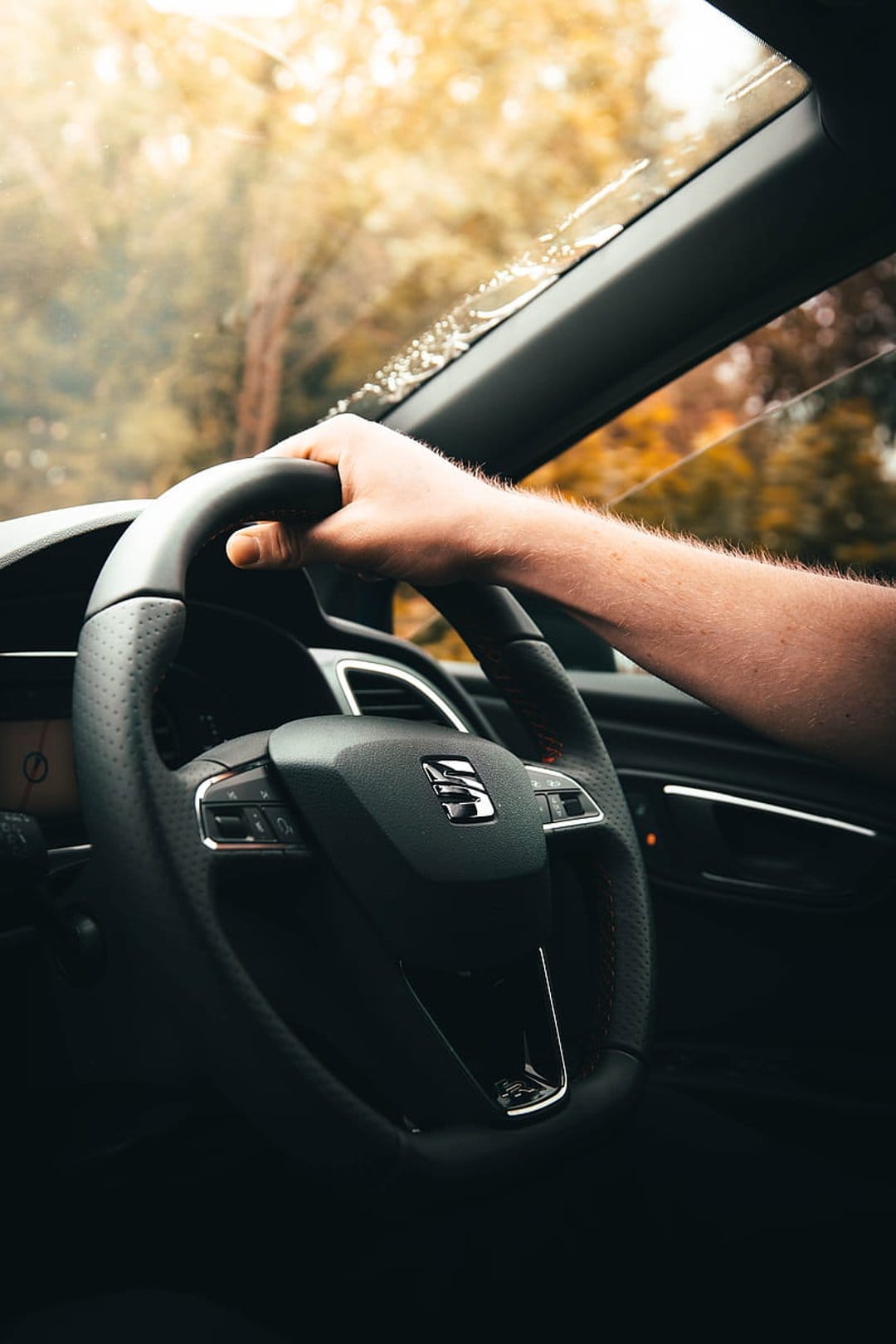 Hand gripping a leather steering wheel inside a car with blurred orange autumn trees visible through the glass.