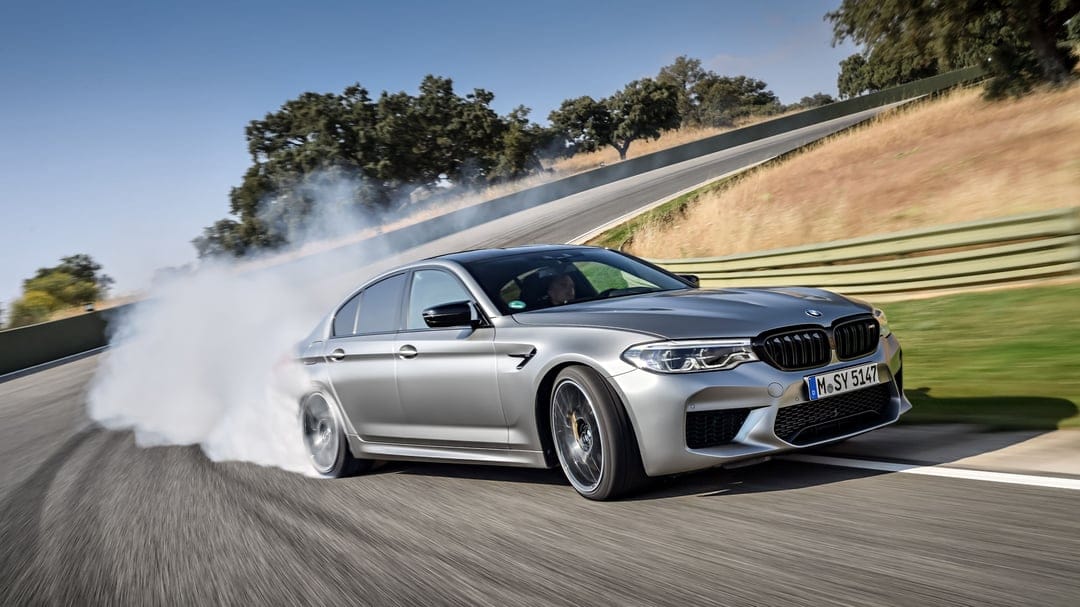 Silver BMW M5 sedan drifting on a paved racetrack with thick white tire smoke and a blurred green landscape.