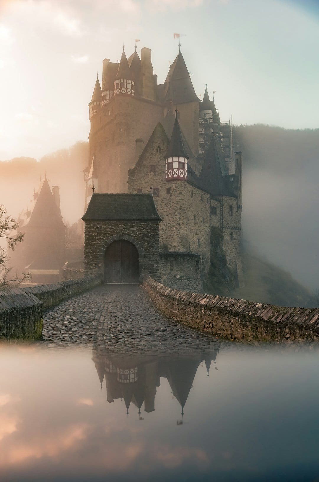 Eltz Castle spires emerge from morning mist with a perfect reflection in still water during golden hour.