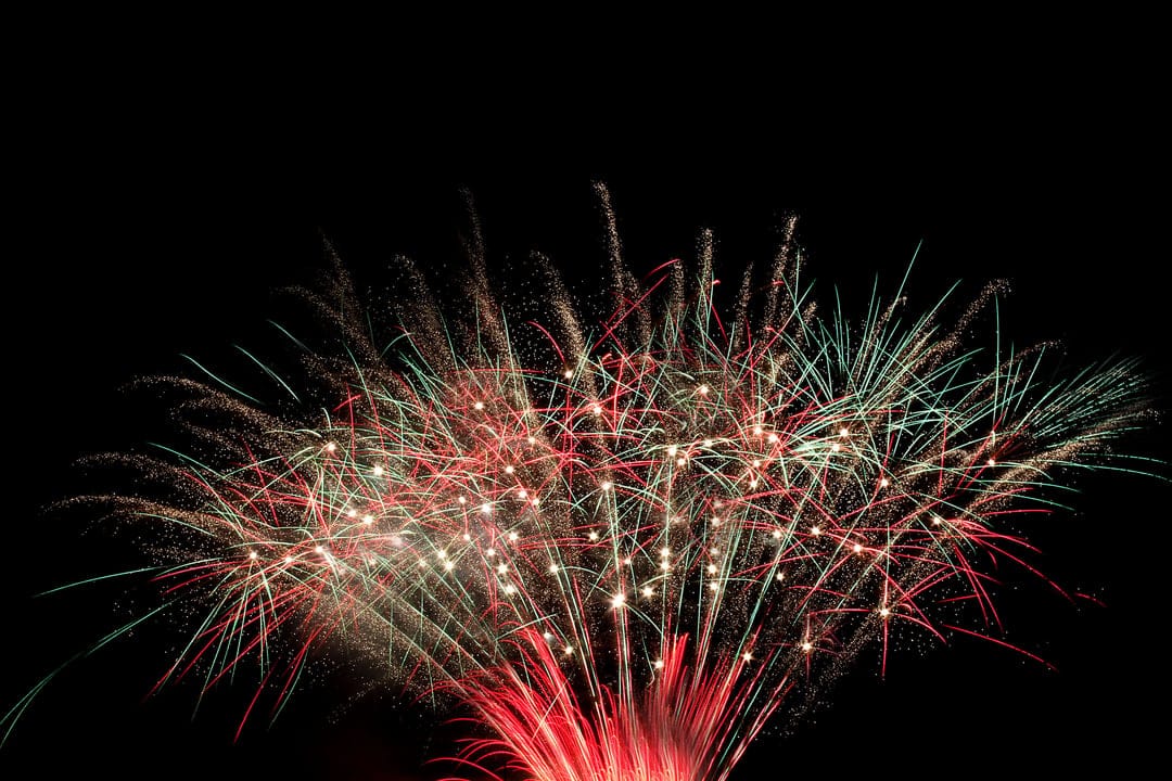 Red and green fireworks burst into luminous streaks against a solid black night sky with radiating light sparks.