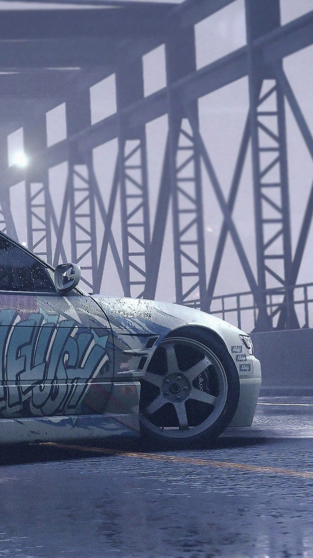Graffiti-covered car parked on a wet street under a metal bridge structure during a foggy, rainy afternoon.