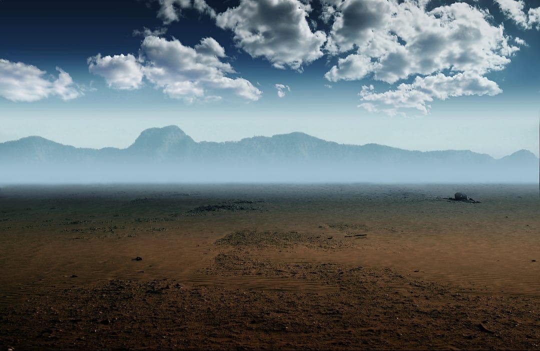 Arid desert valley features textured earth, a dark rock formation, and distant blue mountains under puffy clouds.