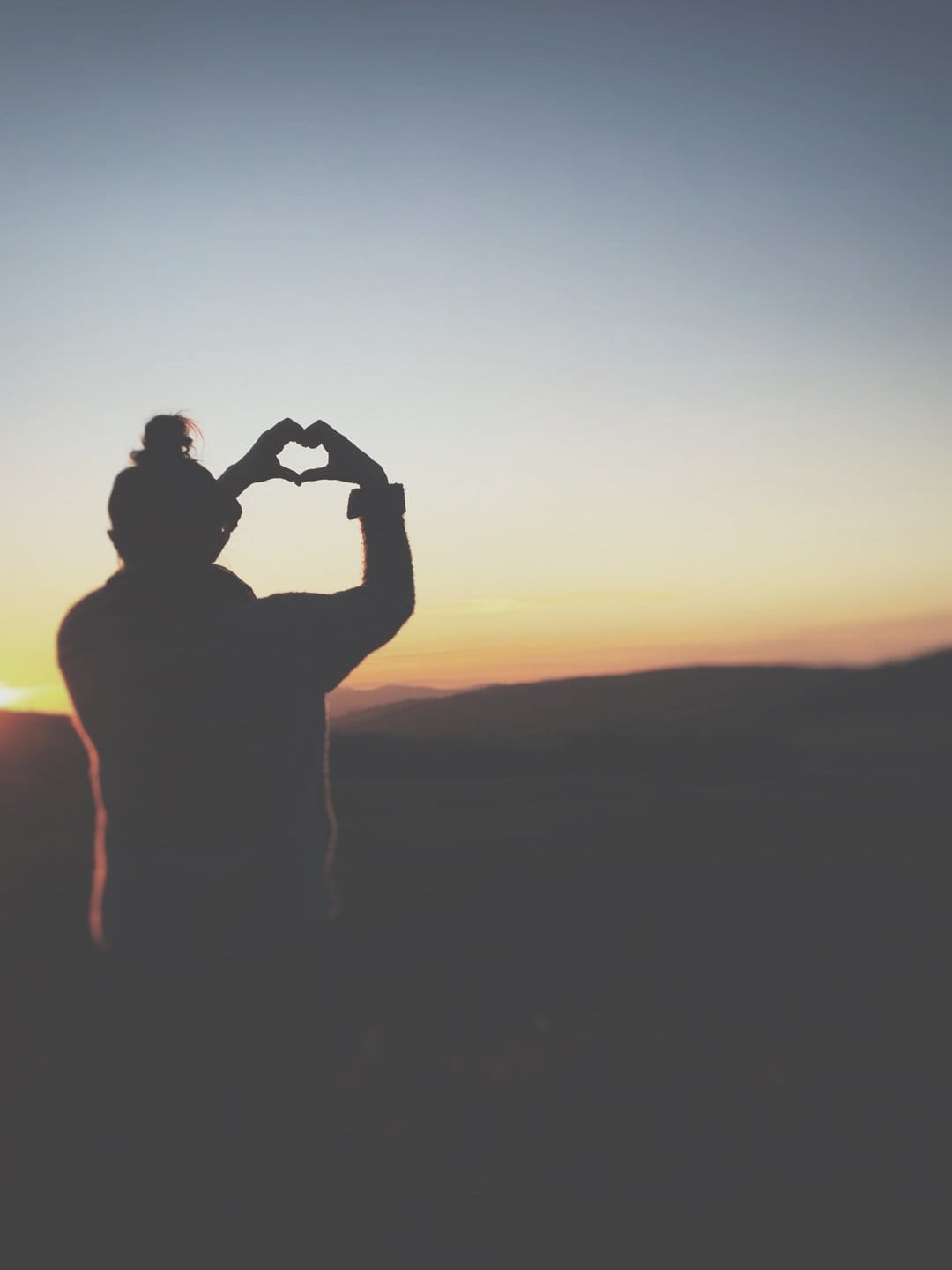 Hand silhouette forming a heart shape over dark mountain ridges during a vibrant orange and blue sunset sky.