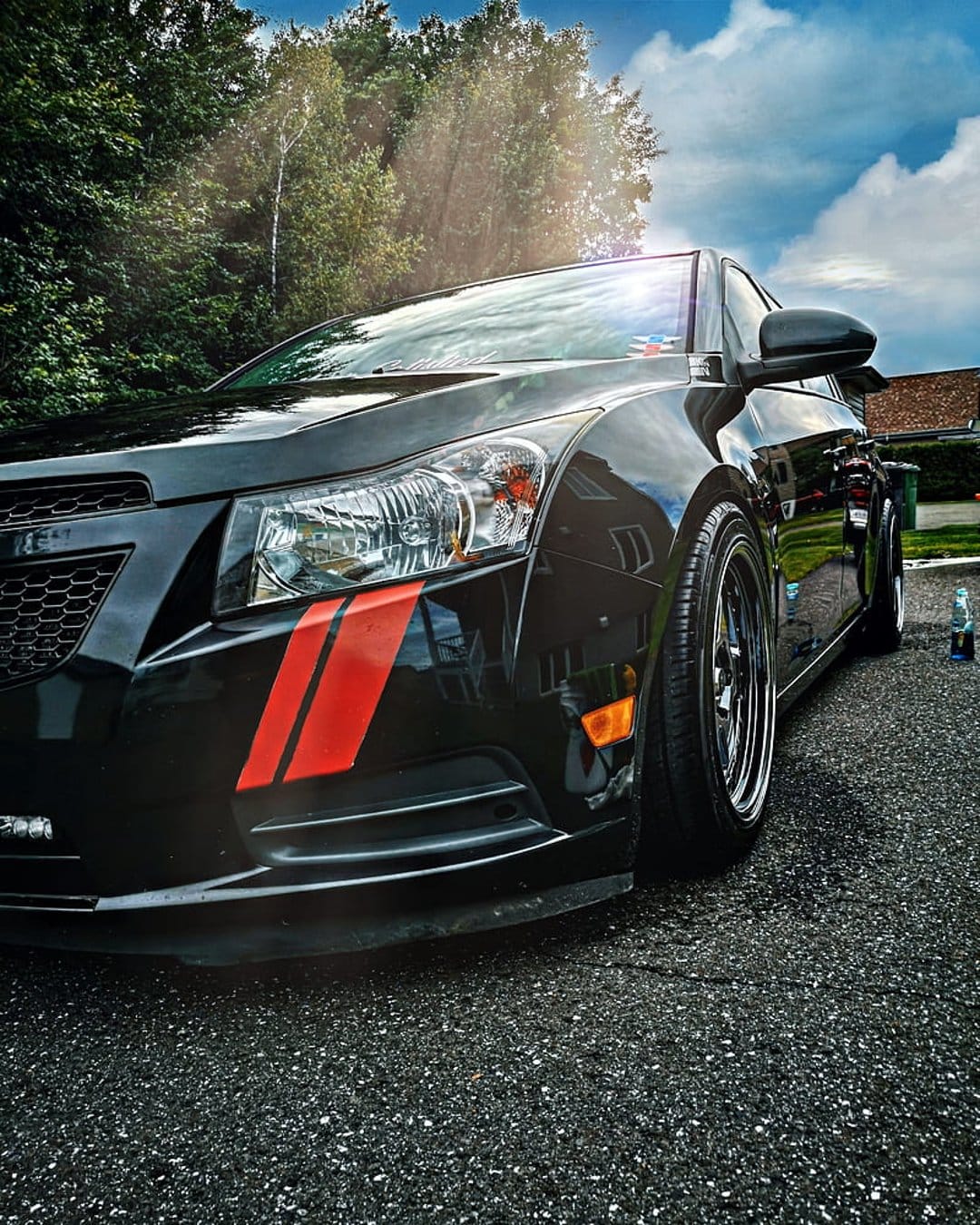 Black Chevrolet Cruze with red fender stripes sits on an asphalt driveway under a cloudy sky and green trees.