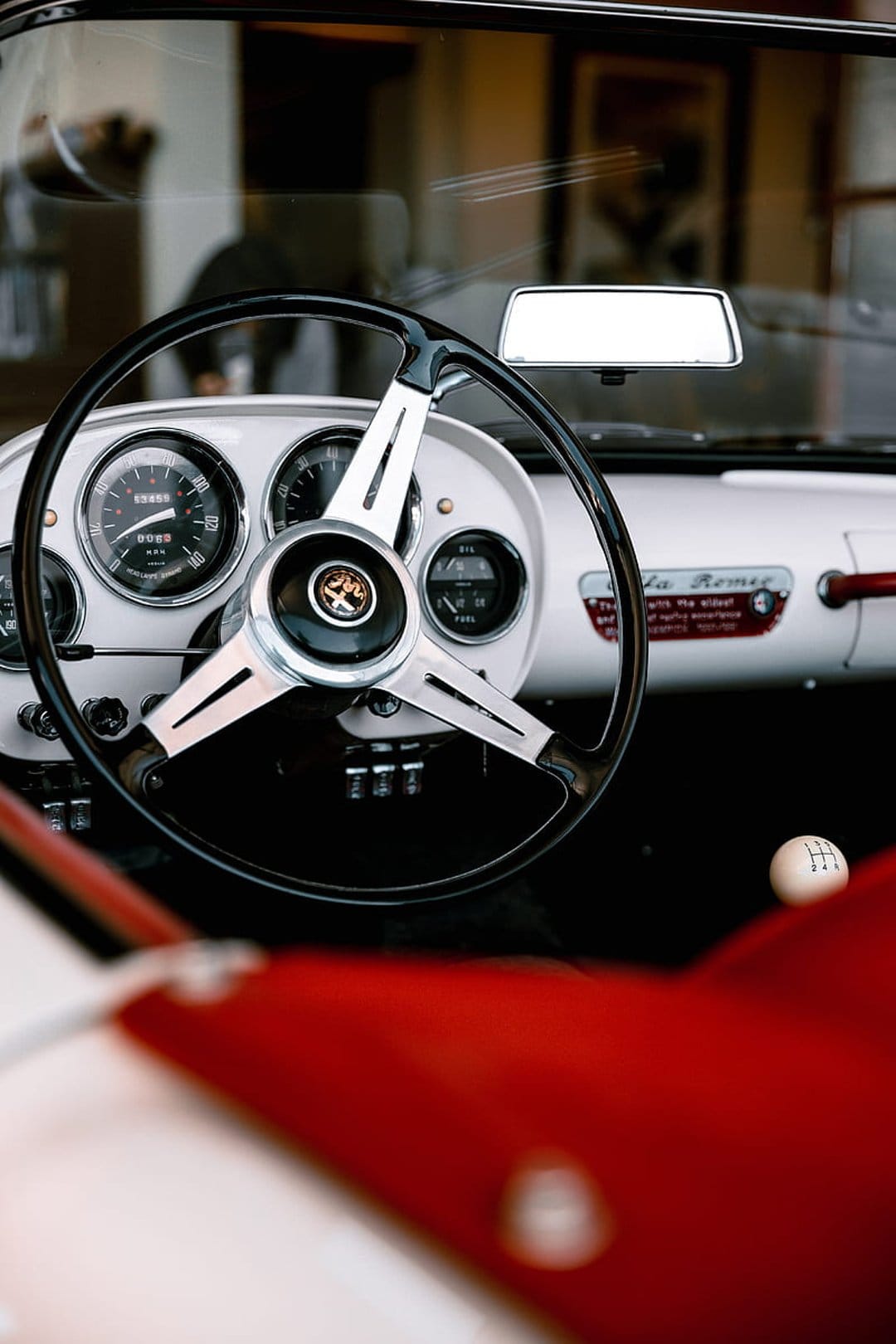 Alfa Romeo steering wheel with chrome horn button, vintage dashboard gauges, and a red leather car seat.