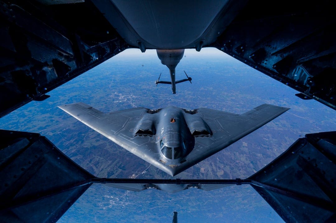 B-2 Spirit stealth bomber receiving fuel from a tanker aircraft boom over a layer of soft white clouds.
