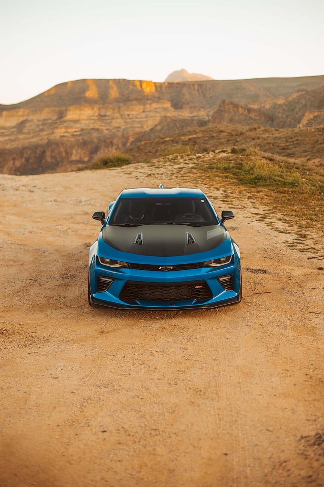 Blue Chevrolet Camaro with a carbon fiber hood parked on a dirt road against sunlit desert hills and canyons.