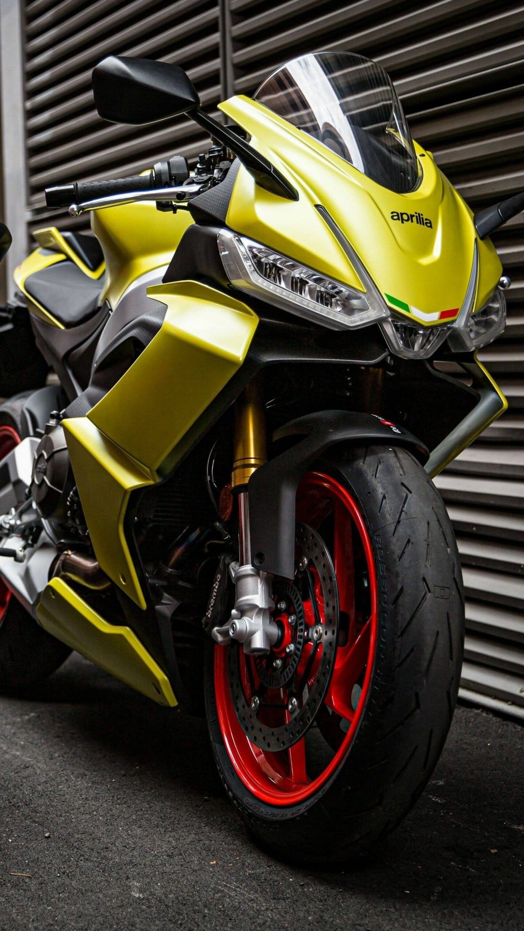 Front view of an acid gold Aprilia motorcycle with red wheels and gold forks against a grey slatted wall.