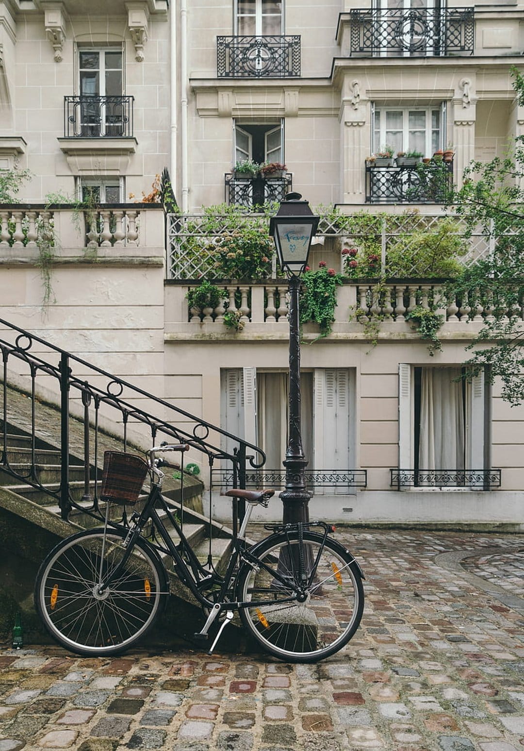 Vintage black bicycle with a wicker basket parked by a stone staircase and a classic Parisian street lamp.