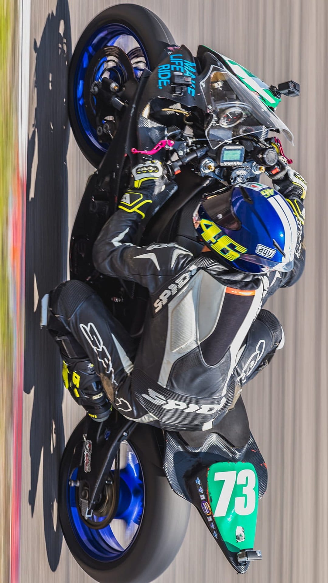 Motorcyclist in a black suit pops a wheelie on a race track with a green number plate and blue helmet.