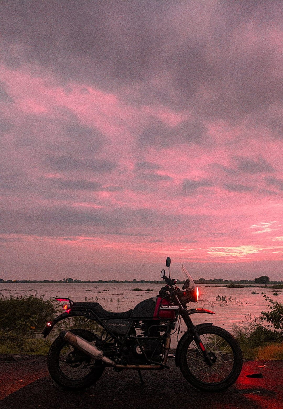 Royal Enfield Himalayan motorcycle parked on a lakeshore beneath a vibrant pink and purple twilight sky.