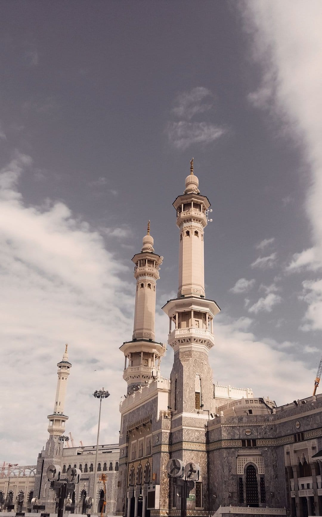 Stone minarets of Masjid Al-Haram stand against a backdrop of thick gray clouds under soft overcast lighting.