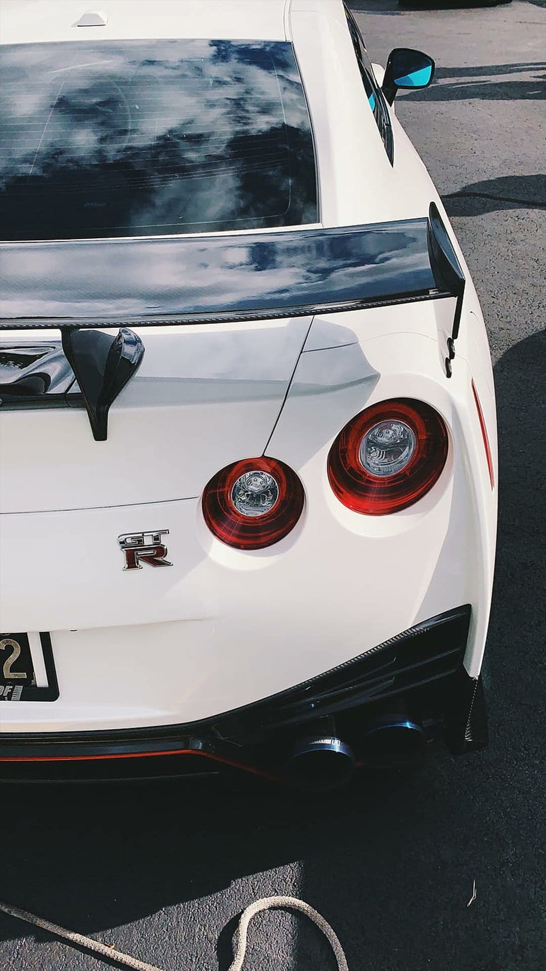 White Nissan GT-R rear view showing four circular taillights, a large spoiler, and a bright blue side mirror.