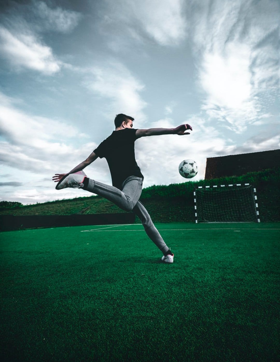 Male footballer kicks a soccer ball mid-air on a green field during a vibrant orange and purple tropical sunset.