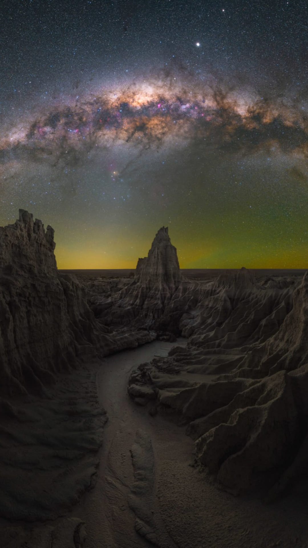 Milky Way galaxy arching over silhouetted desert rock formations and a winding trail under a starry night sky.