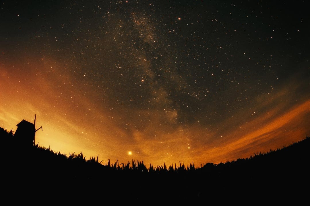 Silhouetted windmill on a hill beneath a vast Milky Way galaxy and a warm orange twilight horizon gradient.