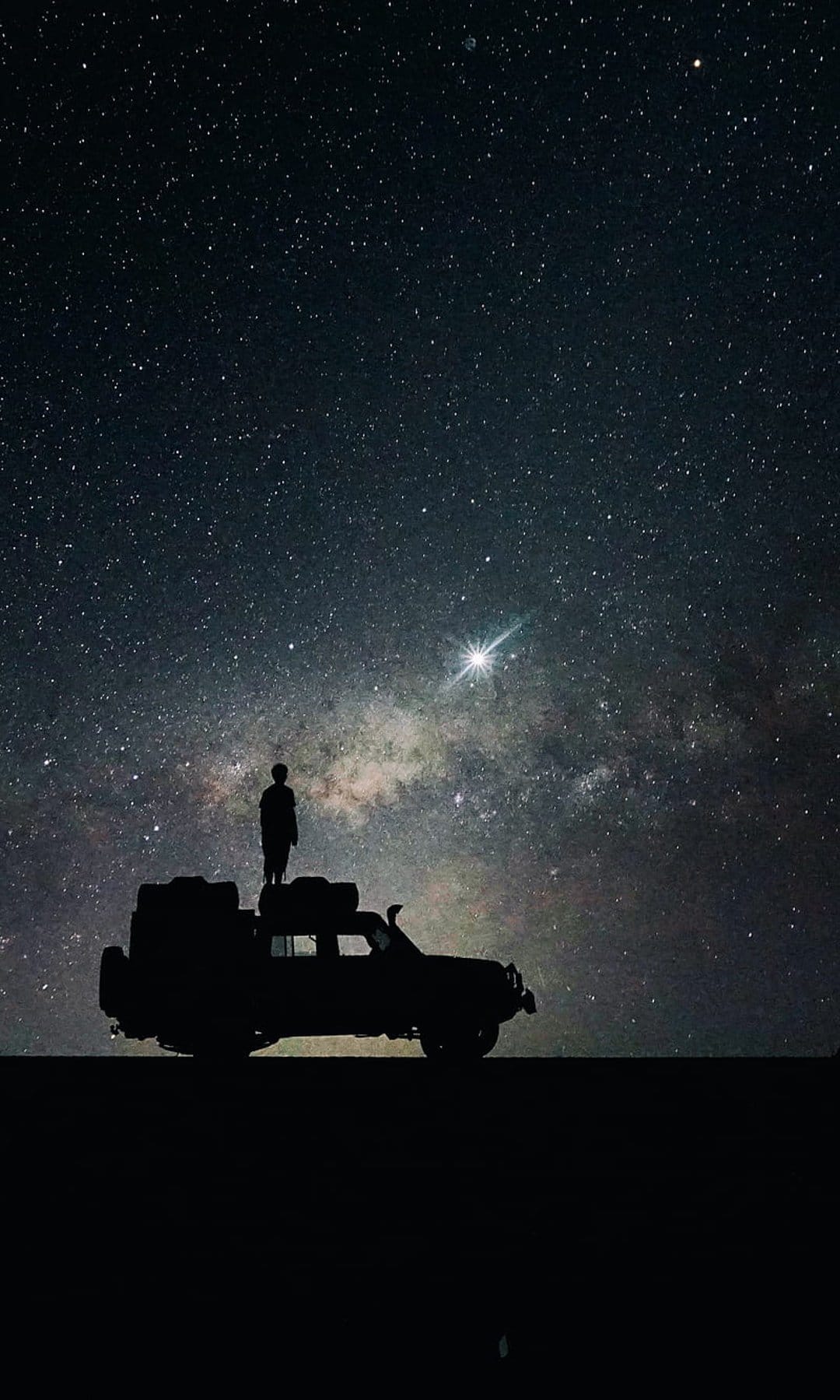 Person standing on an SUV rooftop gazes at the swirling stars and nebulae of the Milky Way in a dark night sky.