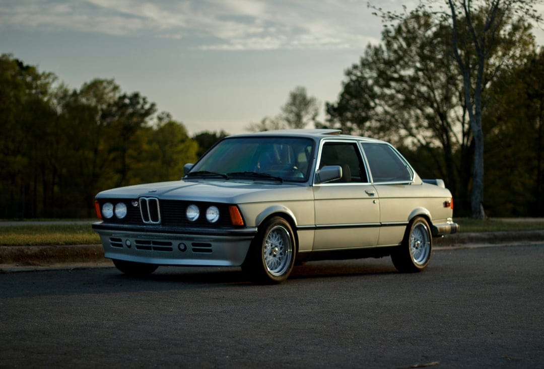 Silver BMW E21 coupe parked on a paved road with blurred green trees and a misty gray sky in the background.