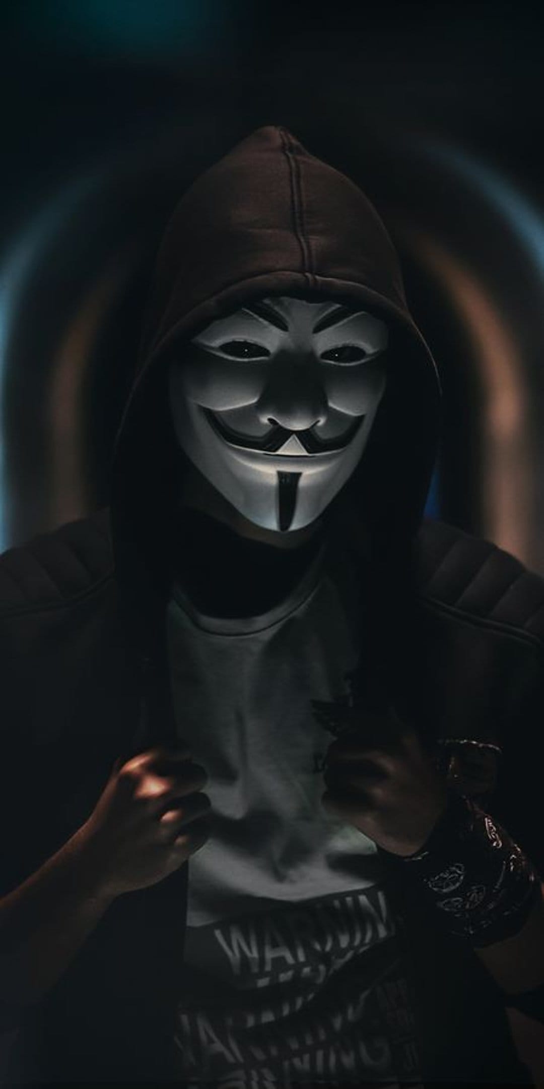Guy Fawkes mask peeks from a dark hood with glowing eyes and deep shadows on a vertical portrait.