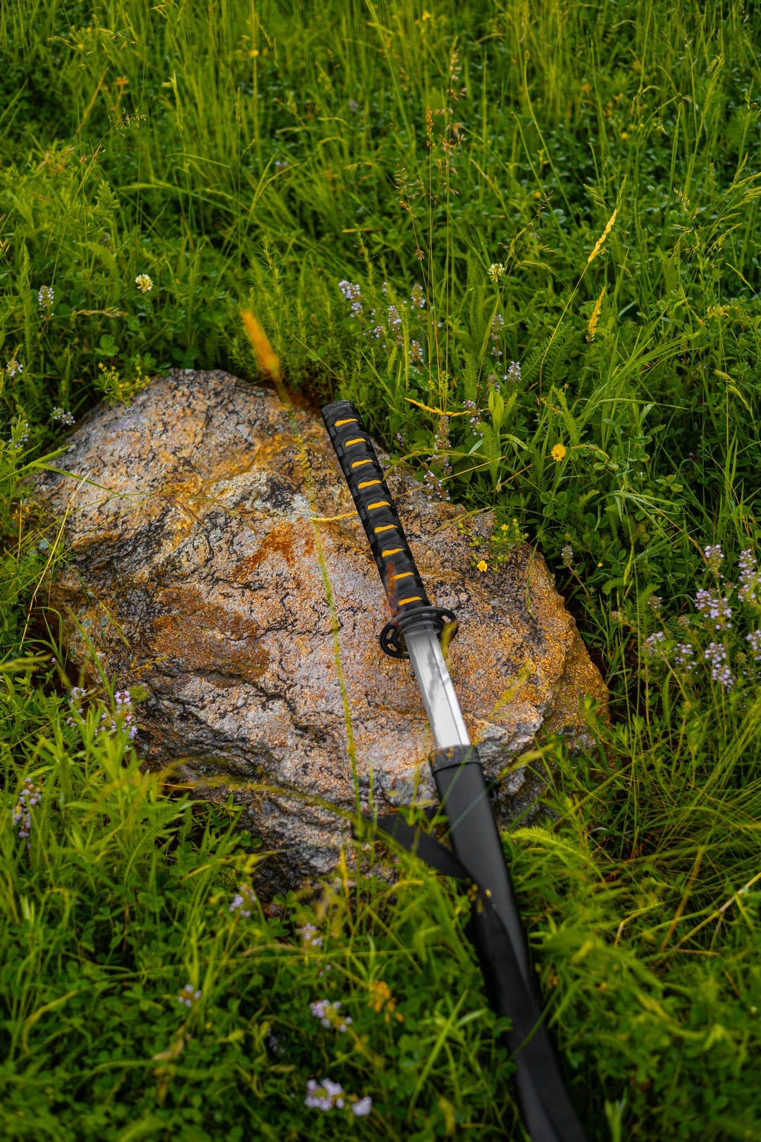 Katana sword with a black handle wrap resting on a grey rock surrounded by green grass and small wildflowers.