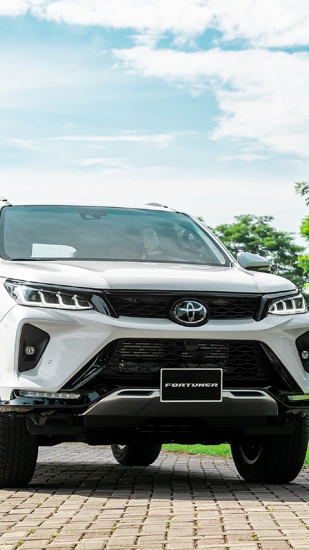 White Toyota Fortuner SUV parked on a brick driveway with headlights and grille detail against a backdrop of trees.