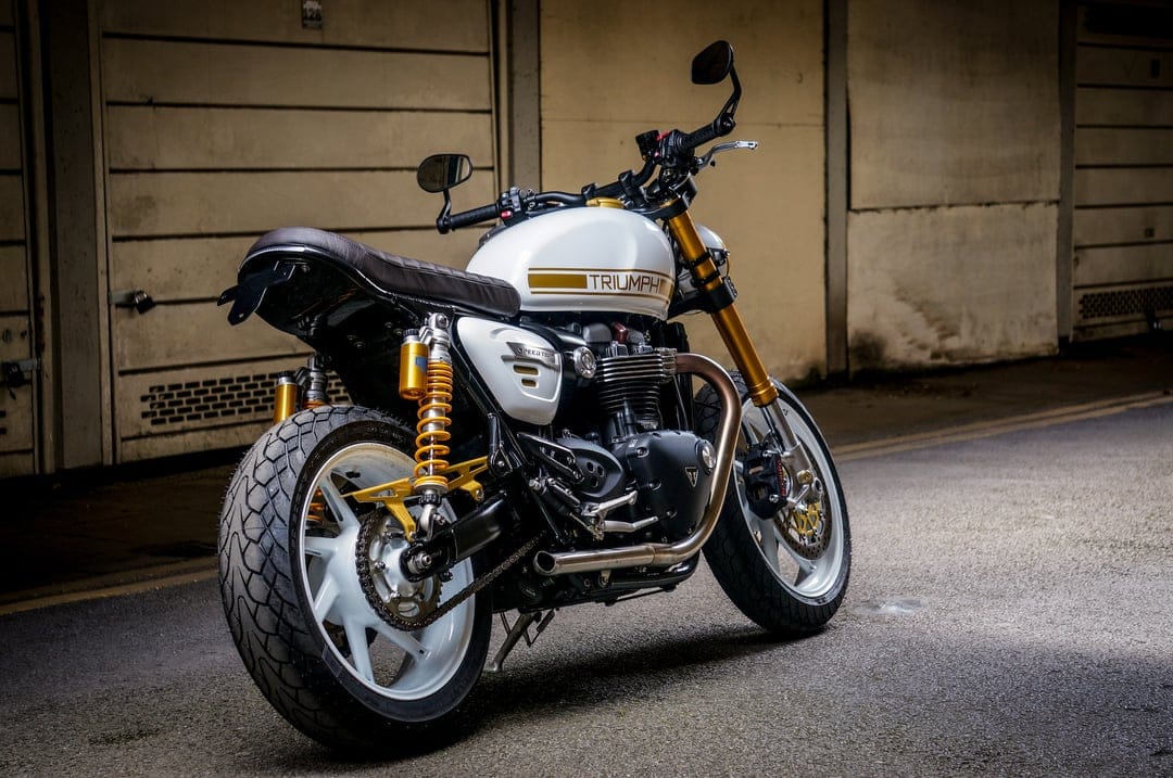 White Triumph Speed Twin motorcycle with gold suspension and white wheels parked in a dimly lit industrial garage.