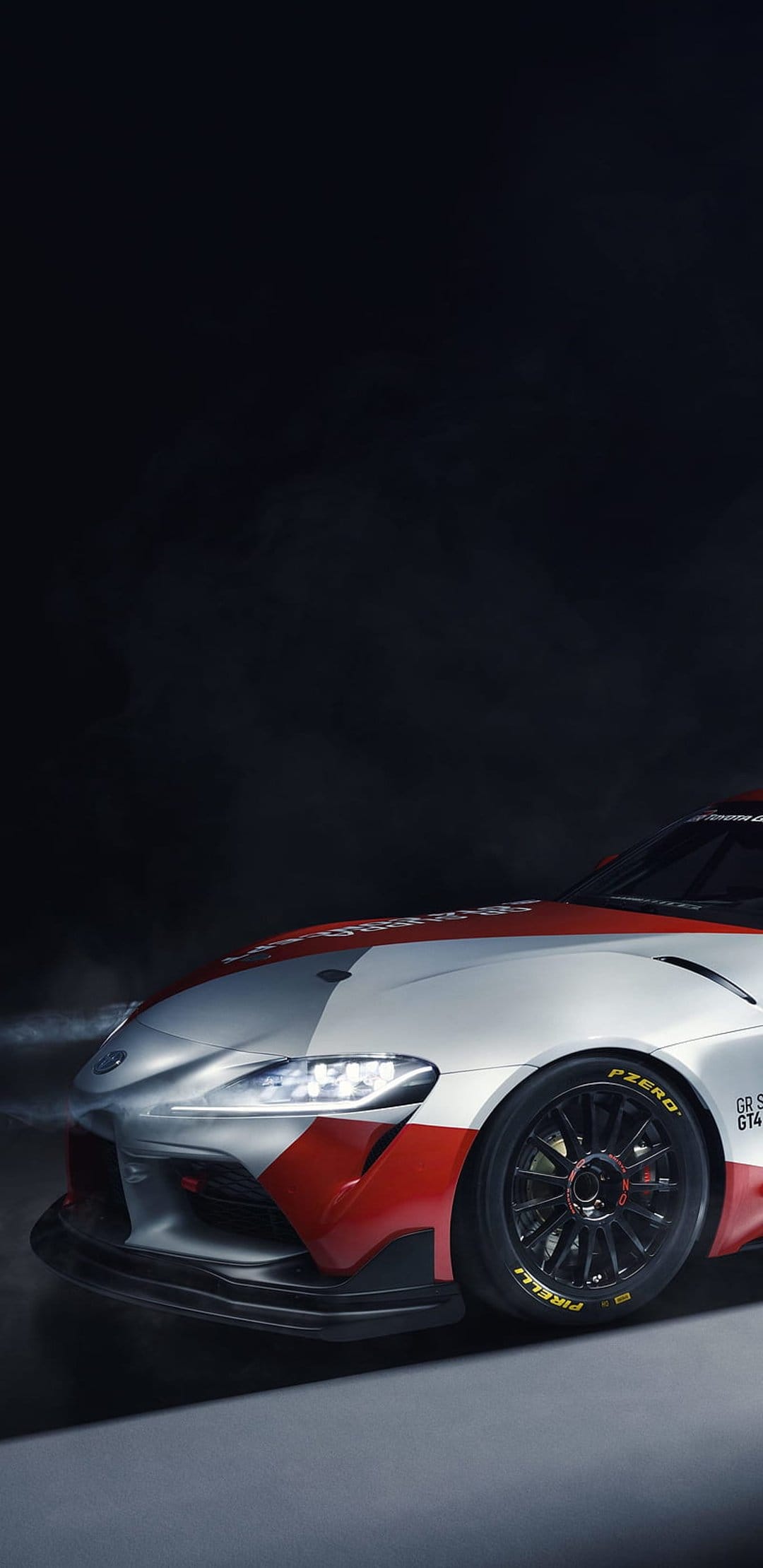 Toyota GR Supra GT4 racing car with glowing LED headlights and Pirelli tires emerges from a dark smoky void.
