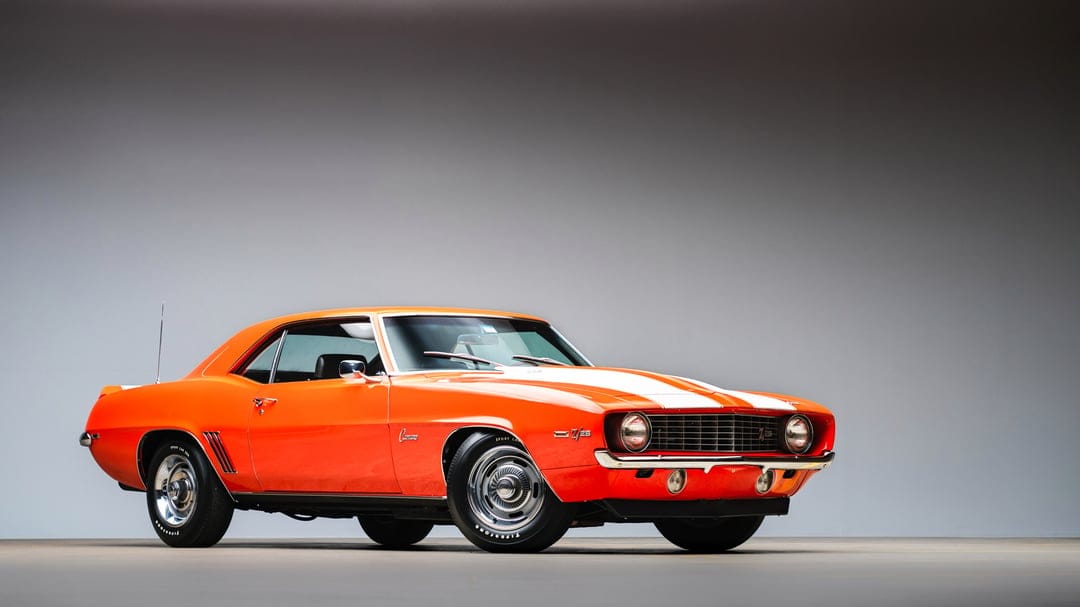 Orange 1969 Chevrolet Camaro Z28 with white racing stripes in a minimalist grey studio setting.