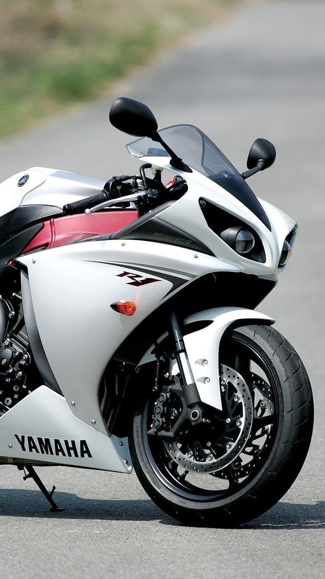 White Yamaha R1 motorcycle parked on asphalt with visible front disc brakes and fairing against blurred trees.