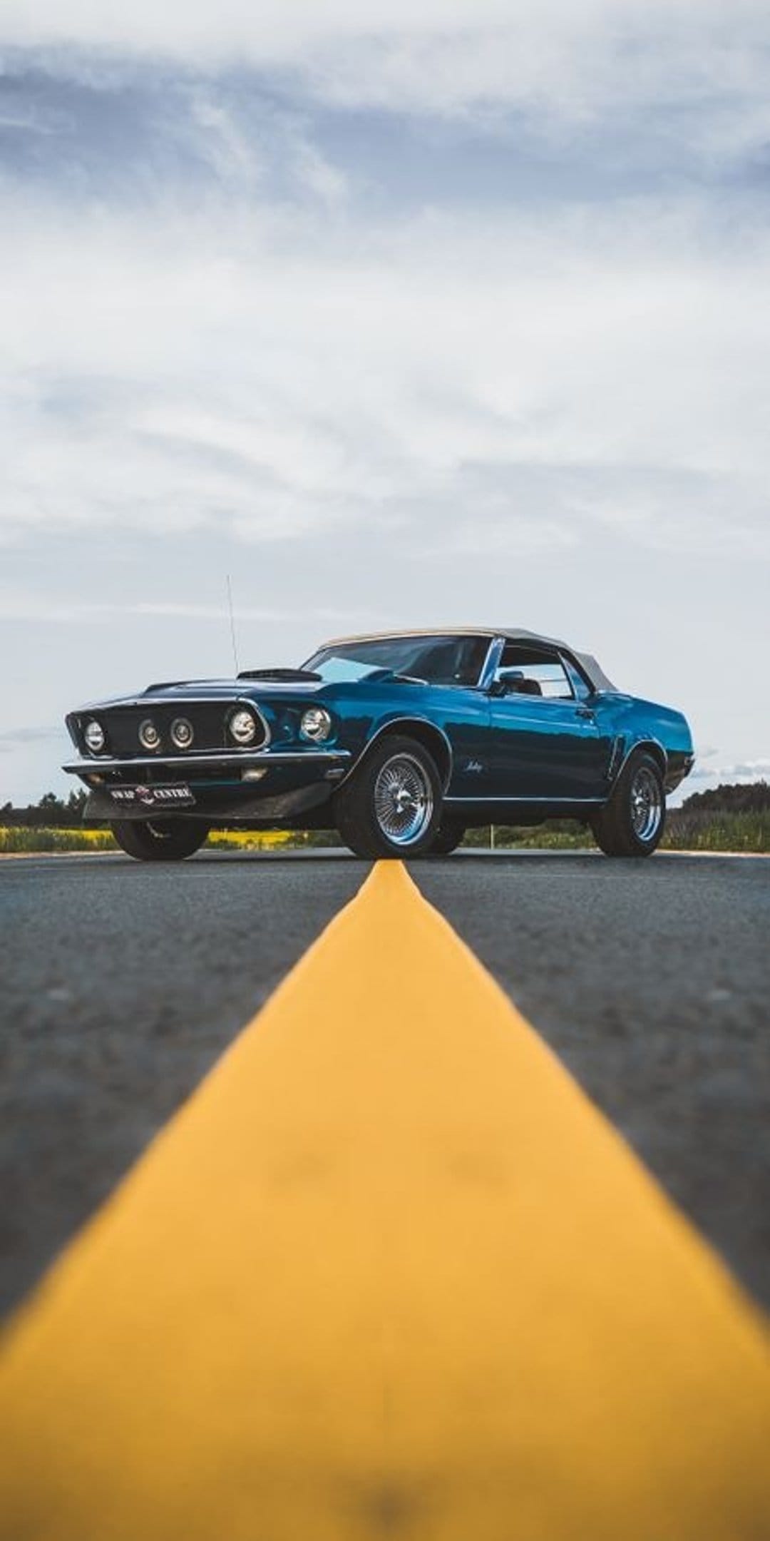 Blue Ford Mustang convertible parked on asphalt with a yellow dividing line under a bright, cloudy daytime sky.