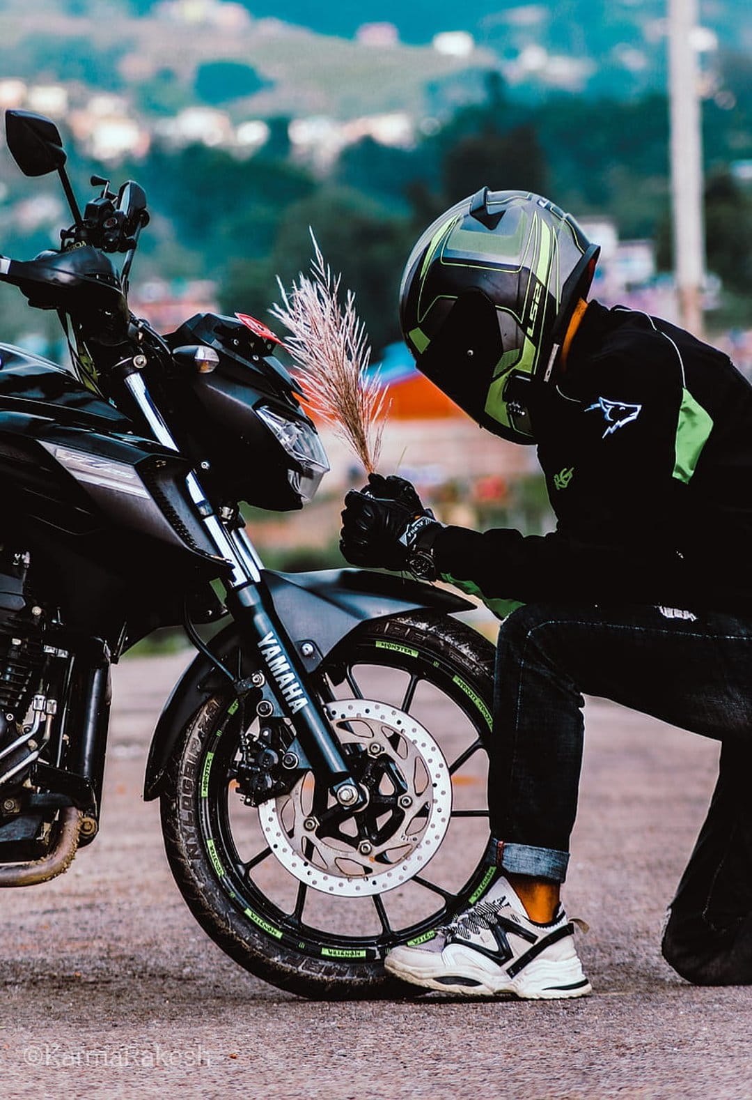 Biker in a green helmet kneels by a black Yamaha motorcycle while holding a plume of dried pampas grass.